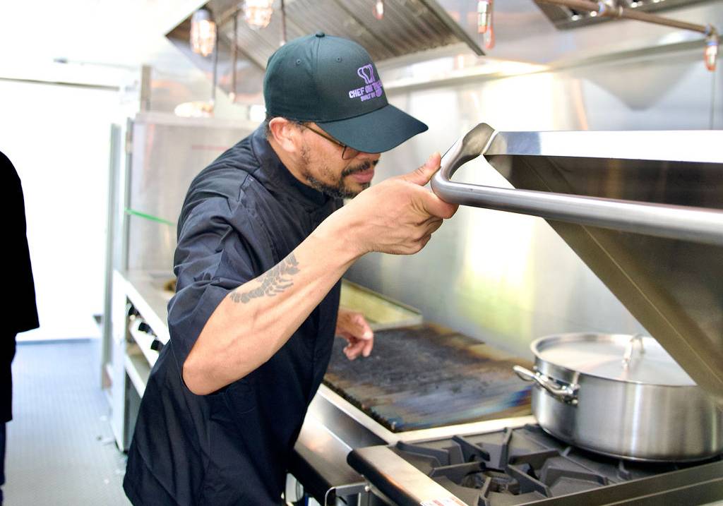 A large white mobile kitchen trailer is parked outdoors on a temporary platform, equipped with ventilation units and propane tanks.
